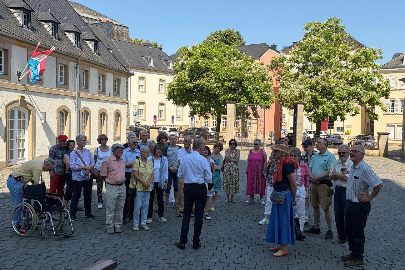 Rektor Erasmy stellt im Schatten der Echternacher Basilika die Architektur vor. Eine Gruppe Teilnehmer hört aufmerksam zu.