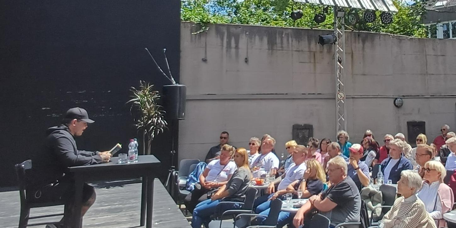 Daniel Schmidt sitzt auf der Bühne an einem Tisch und liest vor einem Publikum von ca. 100 Menschen (c) Thomas Fößel Daniel Schmidt sitzt auf der Bühne an einem Tisch und liest vor einem Publikum von ca. 100 Menschen