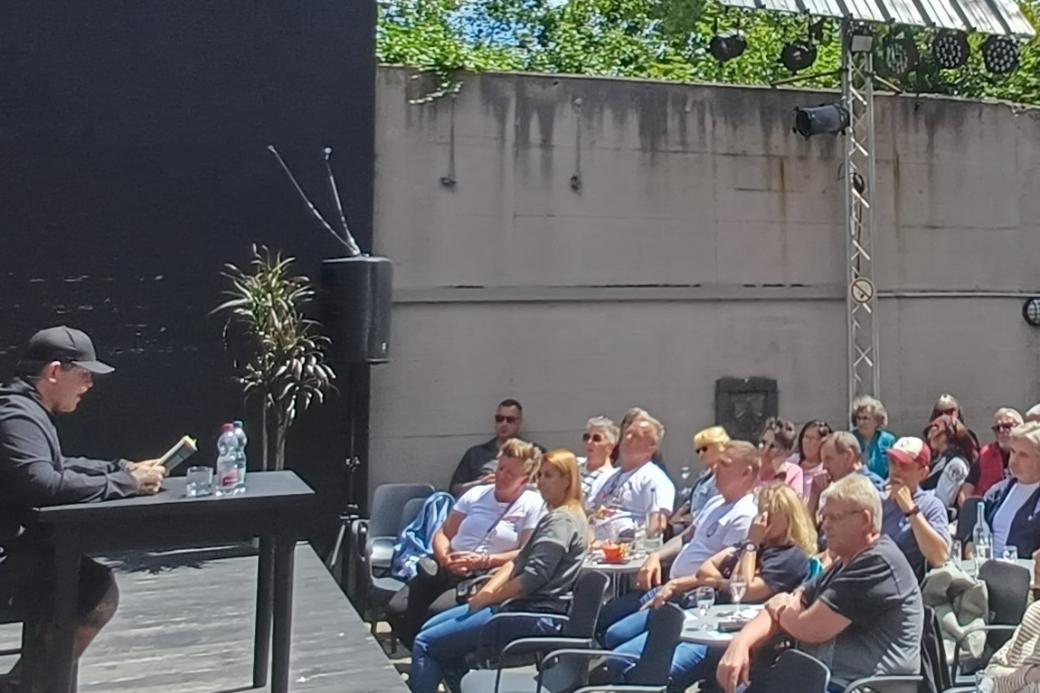 Daniel Schmidt sitzt auf der Bühne an einem Tisch und liest vor einem Publikum von ca. 100 Menschen