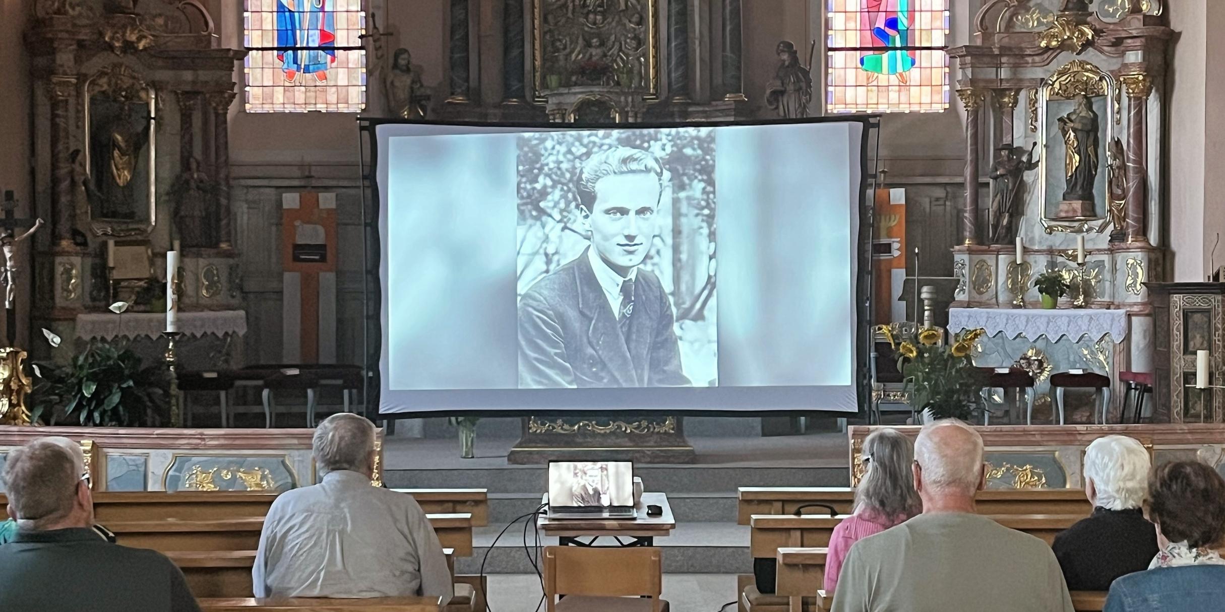 In einer Kirche ist eine Leinwand aufgebaut und Menschen schauen einen Film